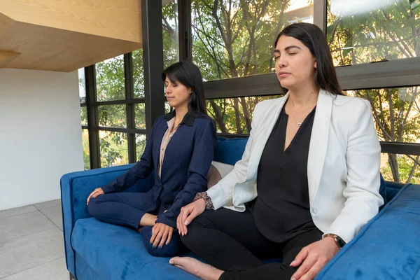 Ofiste çalışırken koltukta meditasyon yapan iki iş kadınının yan görüntüsü.