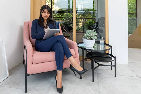 Resmi kıyafetler içindeki genç Latin kadın lobideki koltukta beklerken tablet kullanıyor..