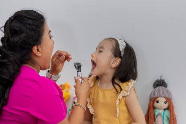 Latin kadın doktor, bir kızın ağzını steteskopla kontrol ediyor.