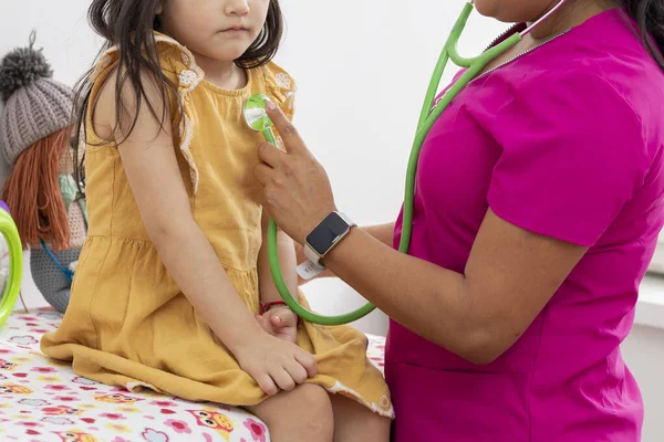 Médico pediatra femenino irreconocible comprobando con un estetoscopio el corazón de una niña con un vestido de color mostaza — Foto de Stock