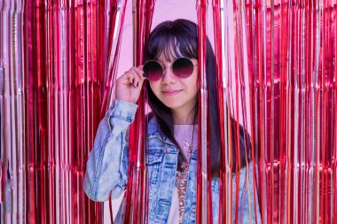 Portrait of teenage girl with glasses behind a metallic pink party curtain.