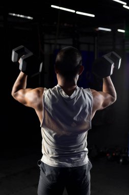 Arkadan bir çift dambılla bastıran kaslı bir adamın dikey görüntüsü.