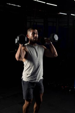 Spor salonunda bir çift dambılla bastıran kaslı bir adamın dikey görüntüsü.