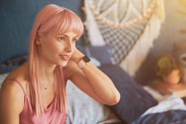 Elegante vrouw met roze haar en moderne smartwatch zit op bed met kussens — Stockfoto