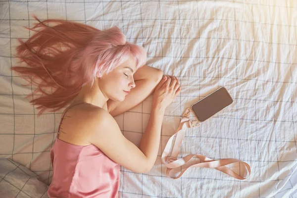 Piękny śpiąca kobieta model z różowymi włosami leży z telefonu komórkowego na dużym łóżku — Zdjęcie stockowe