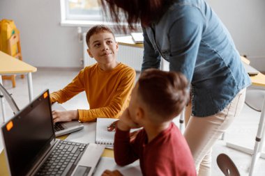 Çocuklar dizüstü bilgisayarda yazıyorlar ve sınıfta öğretmeni dikkatle dinliyorlar. Eğitim, okul kavramı