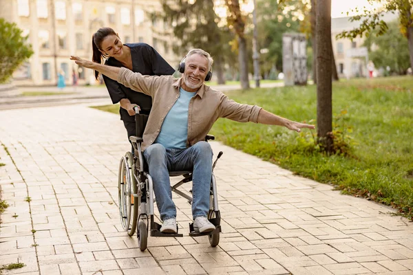 Pria dewasa yang gembira di kursi roda mengenakan headphone bersenang-senang selama berjalan-jalan di kota dibantu oleh perawat muda yang cantik — Stok Foto