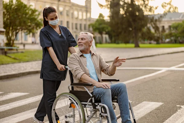 Perawat muda yang peduli dengan topeng wajah yang protektif melakukan percakapan dengan pria cacat di kursi roda saat berjalan-jalan di kota — Stok Foto