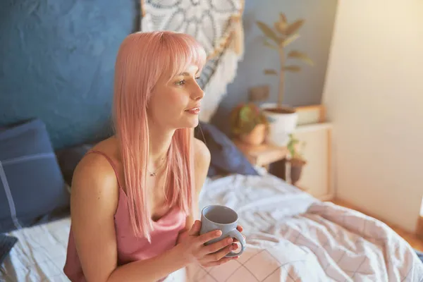 Bedachtzame jonge vrouw houdt beker zittend op comfortabel bed in de ochtend — Stockfoto