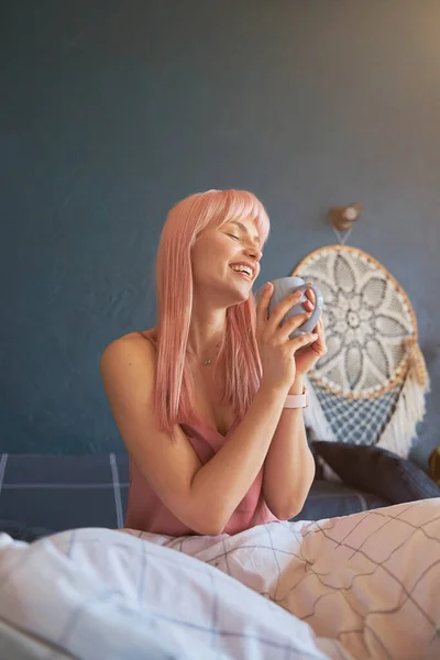 Aangename dame met roze haar houdt mok van heerlijke drank zittend op bed — Stockfoto