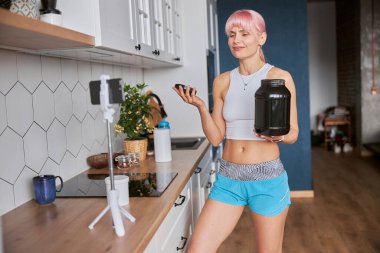 Genç kadın evinde takipçiler için yeni video kaydeden bir kavanoz dolusu beslenme takviyesi tutuyor.