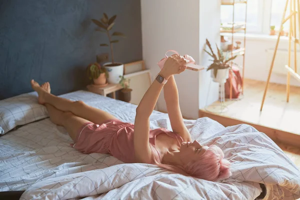 매력적 인 긴 다리를 가진 여자가 큰 침대에 현대의 스마트폰을 들고 셀피를 먹는다 — 스톡 사진