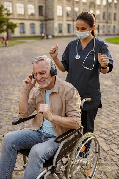 Perawat muda bertopeng pelindung merawat pasien pemulihan senior di kursi roda dengan memakai headphone dan mendengarkan musik di luar ruangan di taman dekat klinik — Stok Foto