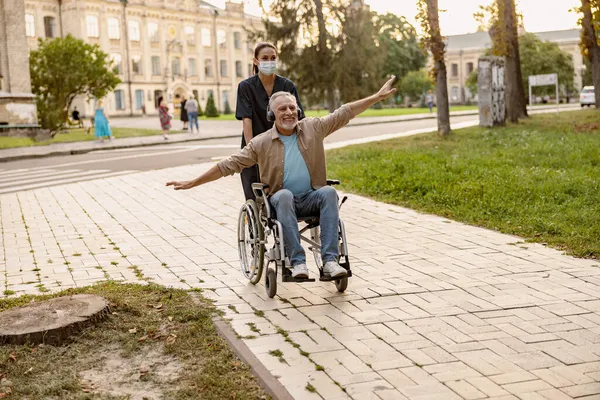 Pria dewasa yang bersemangat di kursi roda mengenakan headphone bersenang-senang selama berjalan-jalan di kota dibantu oleh perawat muda dengan topeng wajah pelindung — Stok Foto