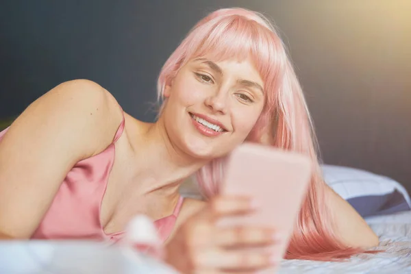 Lächelndes weibliches Model mit rosa Haaren liest Nachricht auf Handy auf bequemem Bett liegend — Stockfoto