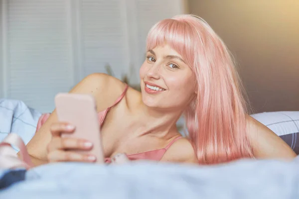 Porträt einer attraktiven Dame mit rosa Haaren, die ein modernes Handy im Bett hält — Stockfoto