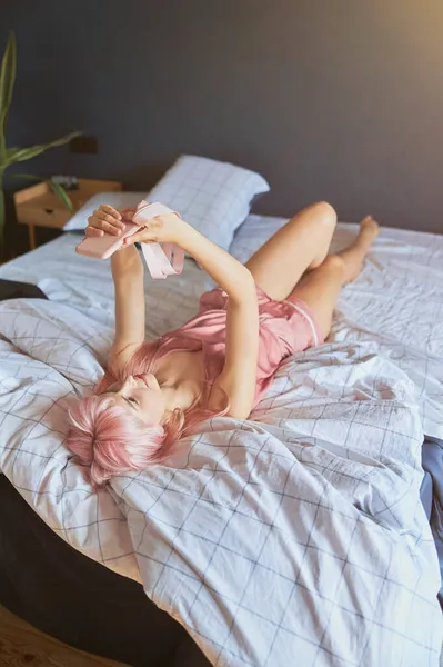 Jeune femme en élégant pyjama de soie prend selfie avec smartphone moderne couché sur le lit — Photo