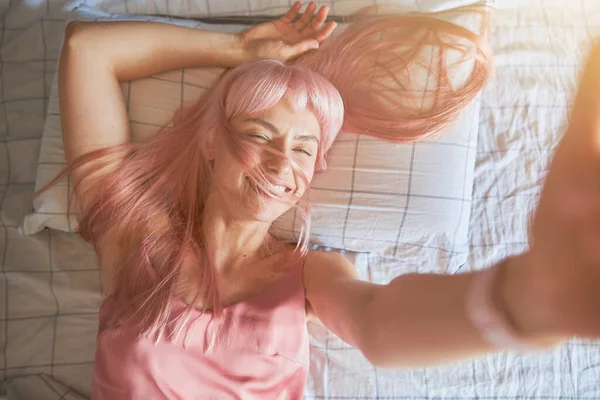 Radosna pani z jasnoróżowymi włosami bierze selfie leżące na wygodnym łóżku — Zdjęcie stockowe