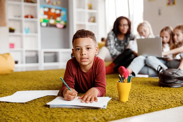 Niño sonriente sosteniendo un lápiz y dibujando sobre papel en el suelo — Foto de Stock