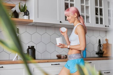 Eşofmanlı neşeli bayan mutfaktaki kameraya beslenme takviyesi kavanozunu gösteriyor.