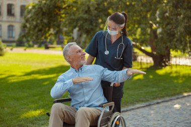 Tekerlekli sandalyedeki neşeli, olgun bir adam yüz koruyucusu ve maskesi takan genç hemşireye gülümsüyor, açık havada yaz günü birlikte yürüyor.