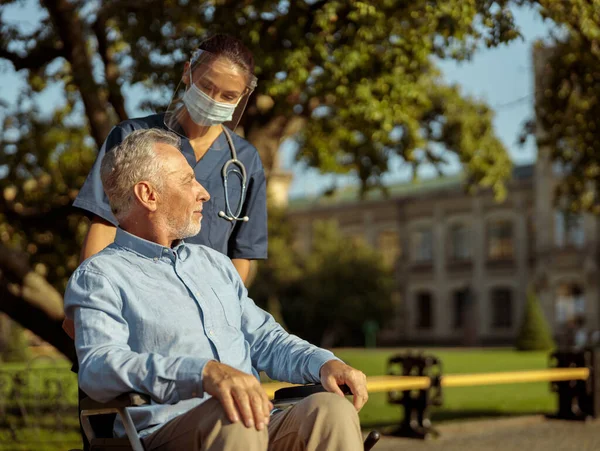 Cuidar a la joven enfermera que usa protector facial y máscara hablando con un hombre maduro, empujando al paciente en silla de ruedas — Foto de Stock