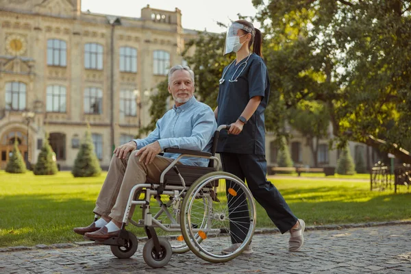 Gambar panjang penuh perawat muda yang peduli mengenakan perisai wajah dan topeng berjalan dengan pasien pria dewasa di kursi roda — Stok Foto