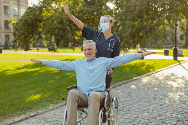 Pria dewasa yang bahagia, memulihkan pasien di kursi roda di jalan-jalan dengan perawat yang mengenakan perisai wajah dan topeng pada hari musim panas di luar ruangan — Stok Foto