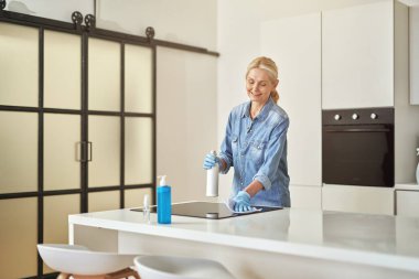 Kauçuk eldivenli gülümseyen olgun ev hanımı gündüz vakti evde bez ve sprey deterjan kullanarak mutfak yüzeyini temizlerken meşgul görünüyor.