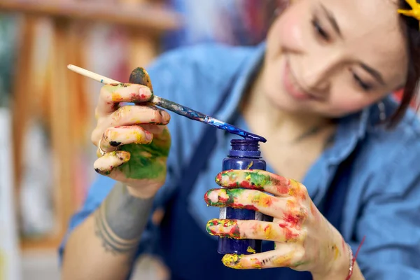 Tersenyum wanita muda di celemek memegang botol cat biru dan kuas cat, bekerja pada sebuah lukisan di bengkel — Stok Foto