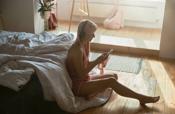 미소짓는 여자는 바닥에 있는 침대 옆에 앉아 헤드폰 과 스마트폰으로 음악을 듣는다 — 스톡 사진