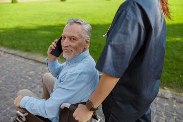 Porträt eines glücklichen älteren Mannes, genesender Patient im Rollstuhl, der in die Kamera lächelt, während er an einem Sommertag im Park mit einer Krankenschwester telefoniert — Stockfoto