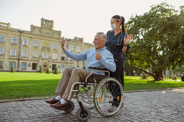 Pria dewasa, memulihkan pasien di kursi roda membuat panggilan video menggunakan smartphone di jalan-jalan dengan perawat mengenakan perisai wajah dan masker — Stok Foto