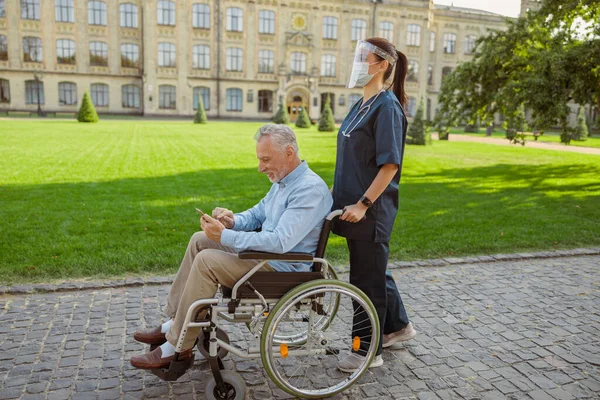 Pandangan samping dari pria senior yang sembuh pasien di kursi roda menggunakan smartphone di jalan-jalan dengan perawat mengenakan perisai wajah dan topeng — Stok Foto