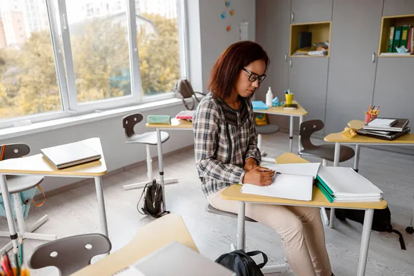 Teacher in glasses sitting at a desk and writing in a notebook — Stock Photo, Image