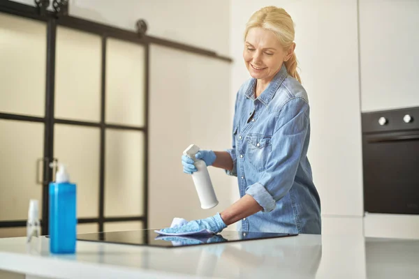 Calm mature blonde woman in rubber gloves cleaning surfaces in the kitchen using cloth and spray detergent at home in the daytime — Stock Photo, Image