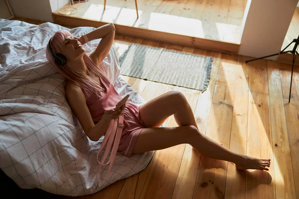 Vrouw met koptelefoon luistert naar favoriete muziek leunend op bed thuis — Stockfoto