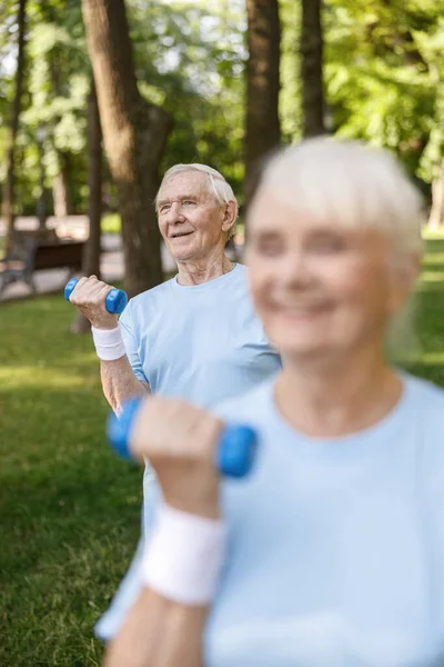Happy senior gentleman does exercises with dumbbell training with wife in picturesque park — Stock Photo, Image
