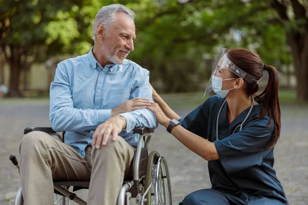 Hombre maduro agradecido, paciente recuperado en silla de ruedas conversando con una enfermera con protector facial y máscara en un paseo por el parque — Foto de Stock