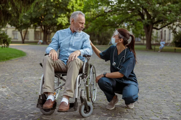 Foto lengkap perawat yang peduli mengenakan pelindung wajah dan topeng berbicara dengan pria dewasa, memulihkan pasien di kursi roda saat berjalan-jalan di taman dekat rumah sakit — Stok Foto