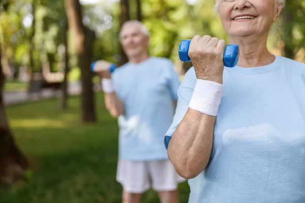 Αθλητική ηλικιωμένη γυναίκα με sweatband κάνει ασκήσεις με dumbbell κατάρτισης με φίλο στο πάρκο — Φωτογραφία Αρχείου