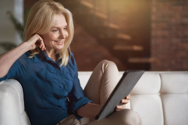 Wanita dewasa tersenyum menonton video menggunakan tablet digital pc, bersantai di sofa di rumah di ruang tamu — Stok Foto