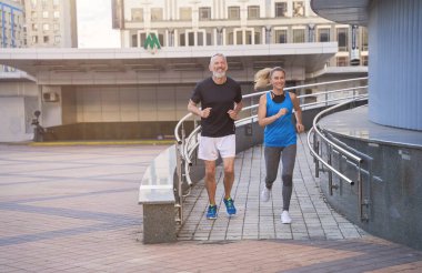 Sportif orta yaşlı çift, spor giyimli erkek ve kadın dışarıda birlikte koşarken mutlu görünüyorlar, kardiyo egzersizi yapıyorlar.