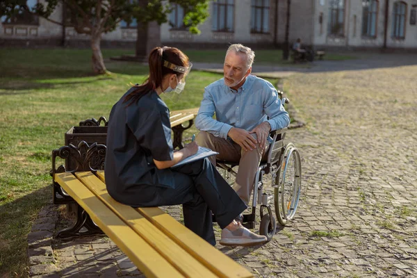Pasien pria di kursi roda yang sedang memulihkan diri berbicara dengan perawat di perisai pelindung wajah di taman dekat klinik rehabilitasi — Stok Foto