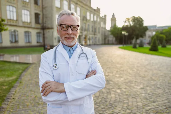 Ritratto di sicuro anziano medico in camice da laboratorio e occhiali sorridenti alla macchina fotografica, in piedi con le braccia incrociate all'aperto vicino alla clinica — Foto Stock