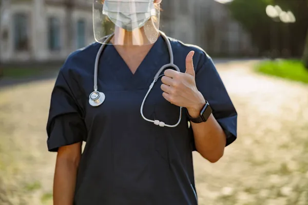 Beskärda skott av ung kvinna sjuksköterska i uniform bär ansikte sköld och mask för säkerhet för att förhindra Covid19, visar tummen upp, står utomhus på en solig dag — Stockfoto