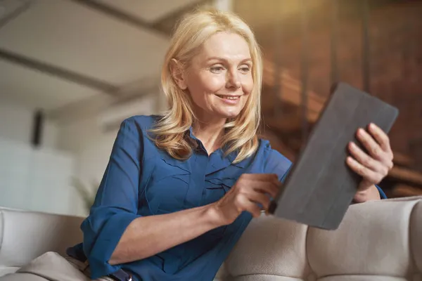 Wanita dewasa yang santai menggulir umpan berita menggunakan tablet digital pc, santai di sofa di rumah di apartemen modern — Stok Foto