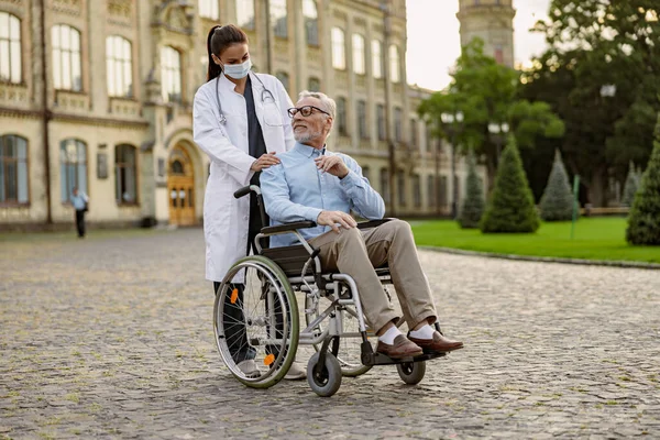 Tembakan panjang dari pria senior pasien cacat di kursi roda tersenyum pada perawat muda dengan topeng pelindung membantunya di luar ruangan — Stok Foto