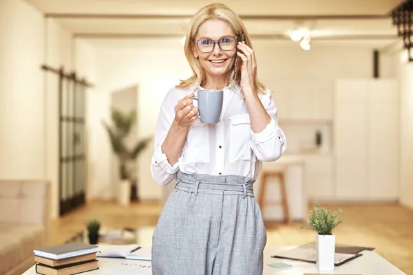 Portrait of atraktif wanita pirang dewasa tersenyum di depan kamera sambil membuat panggilan dan minum kopi, berdiri di kantornya — Stok Foto