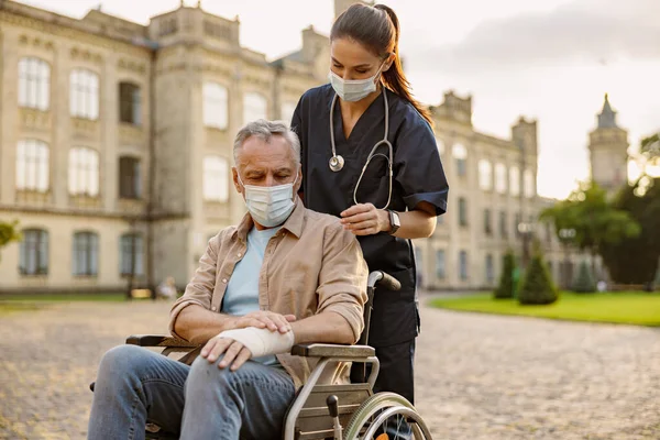 Enfermera joven en máscara protectora cuidando a paciente mayor en recuperación en silla de ruedas con la mano vendada al aire libre en el parque cerca de la clínica — Foto de Stock